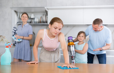 Joint family house cleaning, household help. Daughter clean kitchen table with rag degreasing agent spray. Father teach little female child to vacuum floor, mother wash dish in background blurred