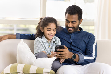Happy pretty little Indian daughter kid and handsome young dad watching funny online content on smartphone, using Internet technology on mobile phone, resting on couch with gadget together
