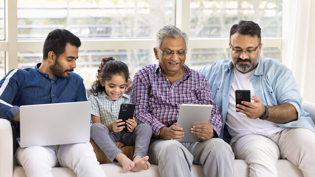 Online addicted Indian kid and men of four generations sitting together on sofa, using application, service, digital gadgets, holding laptop, smartphone, tablet, playing, browsing social media
