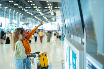 Woman friends looking flight schedule on timetable and go to check-in and boarding gate together in...