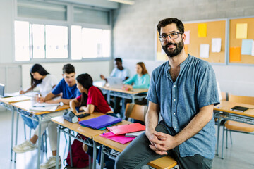 Portrait of young bearded male teacher looking at camera while sitting at high school table over group of diverse students learning together. Education lifestyle concept
