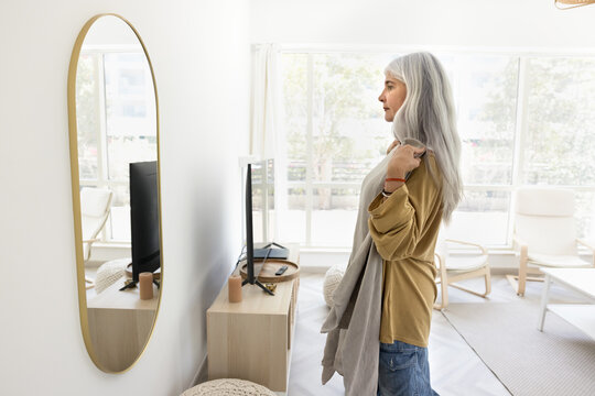 Serious grey haired senior Latin woman trying on cloth at home, looking in mirror. Female fashion webstore customer reviewing purchase, casual grey sweater. Side view