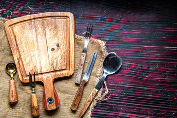 Cutting board, forks, spoons and knives on a dark red wooden table. Space for text.