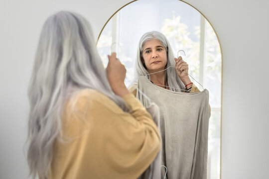 Stylish naturally grey haired senior Hispanic woman choosing oversized sweater for wearing, caring for style, trendy outfit, looking in mirror, trying on cloth on hanger in changing room