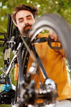 Close-up of sports-loving caucasian man working in his yard to fix bicycle parts with specialized tool. Detailed image of young male cyclist inspecting and adjusting bike wheel and pedal.