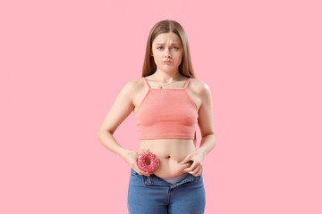 Sad young woman in tight jeans with doughnut on pink background. Weight gain concept