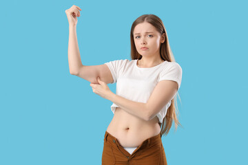 Fototapeta premium Young woman in tight pants with chubby arms on blue background. Weight gain concept