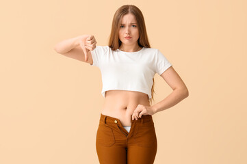 Young woman in tight pants showing thumb-down on beige background. Weight gain concept