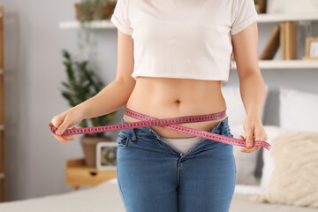Fototapeta premium Young woman in tight jeans measuring her belly at home, closeup. Weight gain concept