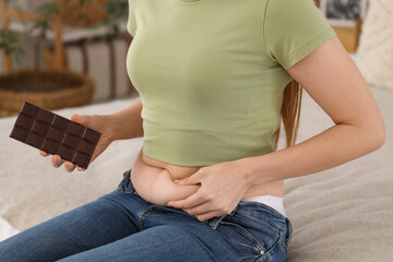 Young woman in tight jeans with chocolate sitting on bed at home, closeup. Weight gain concept