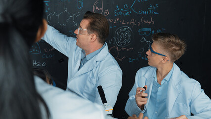 Creative teacher pointing chemistry at blackboard while talking to highschool boy at table with...