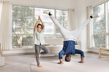 Cheerful Indian yogi dad and little daughter kid doing yoga at home, keeping headstand and tree...