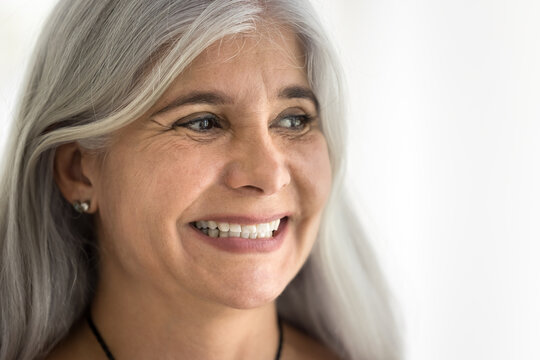 Happy old Latin woman with long grey hair and natural make up looking away with toothy smile. Face of senior female model promoting elderly beauty, dental care. Close up portrait