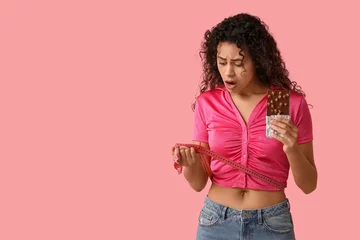 Fotobehang Shocked young African-American woman with sweet chocolate bar and measuring tape on pink background © Pixel-Shot