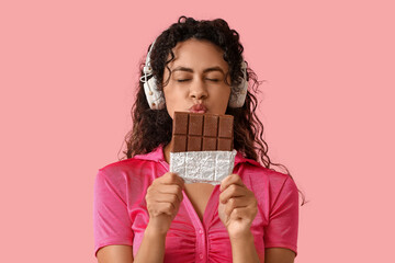 Beautiful young African-American woman in headphones with sweet chocolate bar on pink background