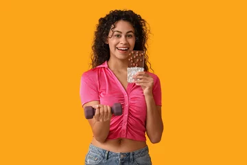 Foto op Plexiglas Happy young African-American woman with sweet chocolate bar and dumbbell on yellow background © Pixel-Shot