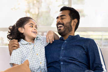 Obraz premium Cheerful caring young Indian dad hugging happy little daughter kid with love, tenderness, smiling, laughing, talking to child, playing, having fun, enjoying fatherhood, family relations
