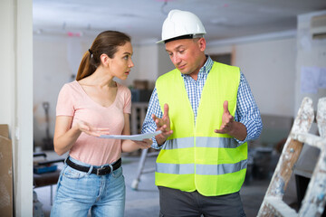 Frustrated young adult female designer arguing with male worker while examining indoor construction...