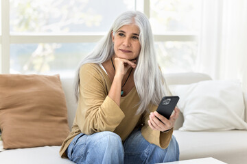 Thoughtful happy elder Latin woman holding smartphone, sitting on home sofa, touching chin, looking away with dreamy smile, thinking, enjoying online communication, Internet technology