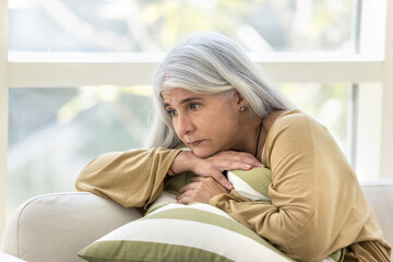 Depressed elderly woman sitting on home couch with sad pointless look away, embracing cushion,...
