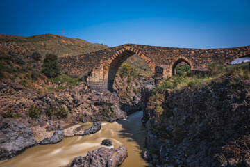 Ponte dei Saraceni. An ancient medieval bridge of Norman age located on the Simeto river. Adrano - Catania, in Sicily. Long exposure picture. June 2023