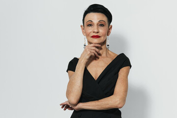 Elegant senior woman in black dress and earrings striking a pose with hand on chin before camera