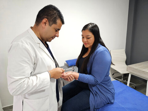 Dark-skinned male doctor attends to Latin woman patient neurological examination with reflex hammer using percussion on tendon