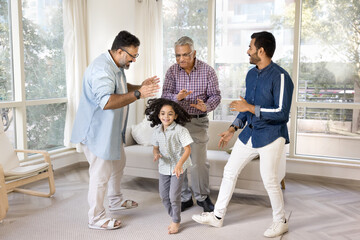Happy Indian men of three family generations and little kid dancing at home together, celebrating family event. Girl having fun, enjoying motion, activity with dad, granddad and great grandpa