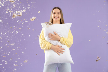 Young woman with soft pillow and flying feathers on lilac background