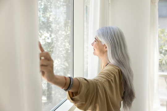 Happy inspired senior Latin woman opening white transparent veils, enjoying morning daylight, looking out of large window, smiling, relaxing in comfortable apartment. Side view,