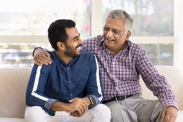 Cheerful elderly Indian granddad and happy young adult grandson talking on home couch, hugging,...