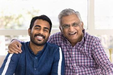 Cheerful elderly Indian father hugging young adult son, touching shoulder with support, parents pride, looking at camera with toothy smiles, laughing, having fun, posing for head shot portrait