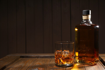 Whiskey with ice cubes in glass and bottle on wooden crate against black background, space for text