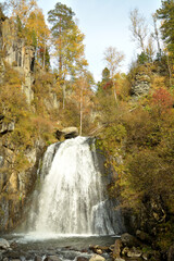 The rapid flow of the river flows like a stormy and powerful waterfall from a high mountain surrounded by an autumn forest.