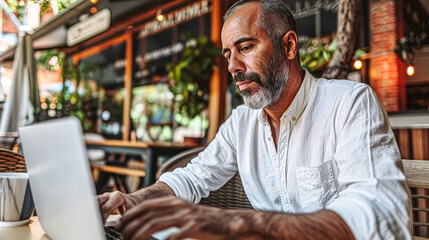 A man is sitting at a table with a laptop in front of him. He is wearing a white shirt and he is focused on his work. The scene takes place in a restaurant or cafe, with a potted plant nearby - Powered by Adobe