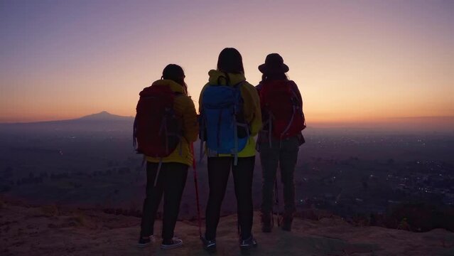 Three female hikers with hiking backpacks and yellow jackets reach the top of the mountain, reflecting friendship and adventure in nature on a bright day.