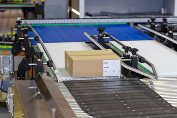 Fototapeta premium Logistics Efficiency - Cardboard Box on Conveyor Belt in Distribution Warehouse. Parcel Moving Through Automated Shipping System. Streamlined Order Fulfillment - Box in Transit on Warehouse Conveyor