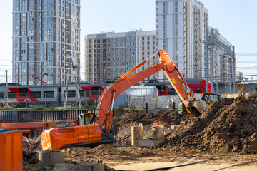 A new orange excavator is working on a construction site with a pile of brown soil. High-rise...