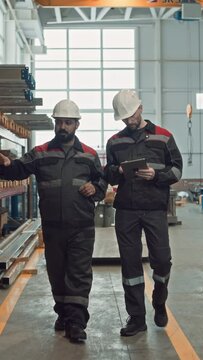 Vertical full shot of two multiracial male workers in coveralls and hard hats walking along industrial factory while inspecting machinery equipment and talkingVertical full shot of two multiracial mal