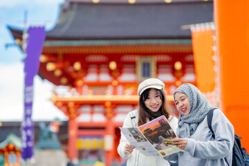 Travel, muslim, Two Asian female tourists of different religions friends visitor learning about history of fushimi inari shrine in travel book while walking through senbon torii path in Kyoto Japan.