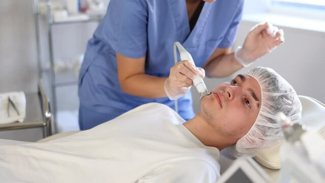 Young female cosmetologist performs hardware facial microcurrent procedure to young male patient