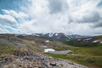 Fototapeta premium Beautiful small lake with snowfield among green grassy hills and rocks with view to snow-capped mountain range. Scenic landscape with alpine lake and snowy high mountains. Mountain lake in highlands.