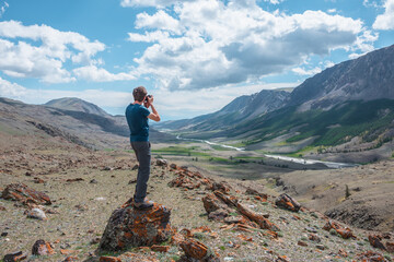 Man shoots landscape with mountain river in alpine valley. Guy with photo camera on stone with view...