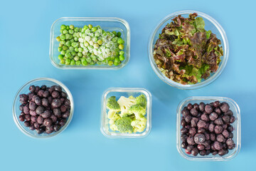 Green vegetables and berries in glass lunch boxes on blue background close-up, top view.