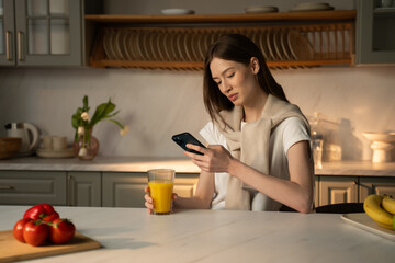 A serene scene unfolds as a young woman, casually dressed, stands at a kitchen counter bathed in soft morning light. She smiles gently while engaging with her smartphone, possibly texting or browsing