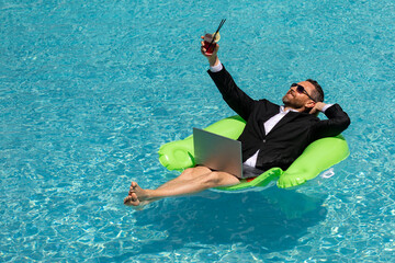 Funny businessman in suit working with computer on the sea beach. Summer holiday, outdoor and business travel concept. Crazy office worker. Man working on laptop computer sitting in cloth at poolside. - Powered by Adobe