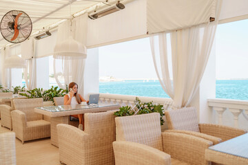 Woman coffee cafe laptop sea. Female freelancer using laptop on coffee break in modern cafe with sea view in the morning