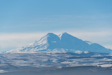 View from the road of the snow-covered majestic double-headed Elbrus in the morning haze. Close-up. Elbrus is the main attraction of Kabardino-Balkaria. December 2023