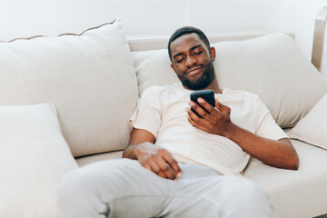 Happy African American man sitting on a black sofa in his modern apartment, typing on his mobile...