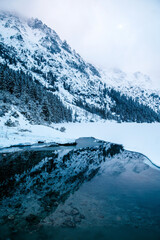 Morskie Oko lake sits in front of a majestic snowy mountain range, creating a stunning natural landscape. The sky is filled with fluffy clouds, enhancing the atmosphere of the scene. Tatry, Poland